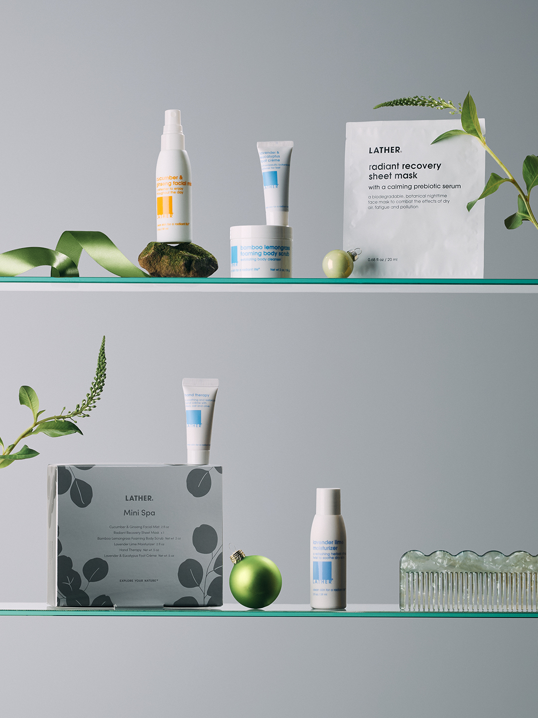 LATHER Mini Spa gift box and hand therapy cream on the lower shelf, with cucumber & ginseng facial mist, lavender & eucalyptus foot crème, and a radiant recovery sheet mask on the shelf above, styled with greenery and a green ornament.