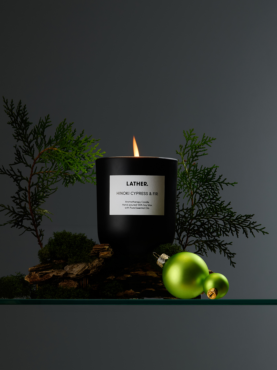 A lit LATHER Hinoki Cypress & Fir candle resting on bark and moss, flanked by cypress branches and green ornaments against a dark grey background.