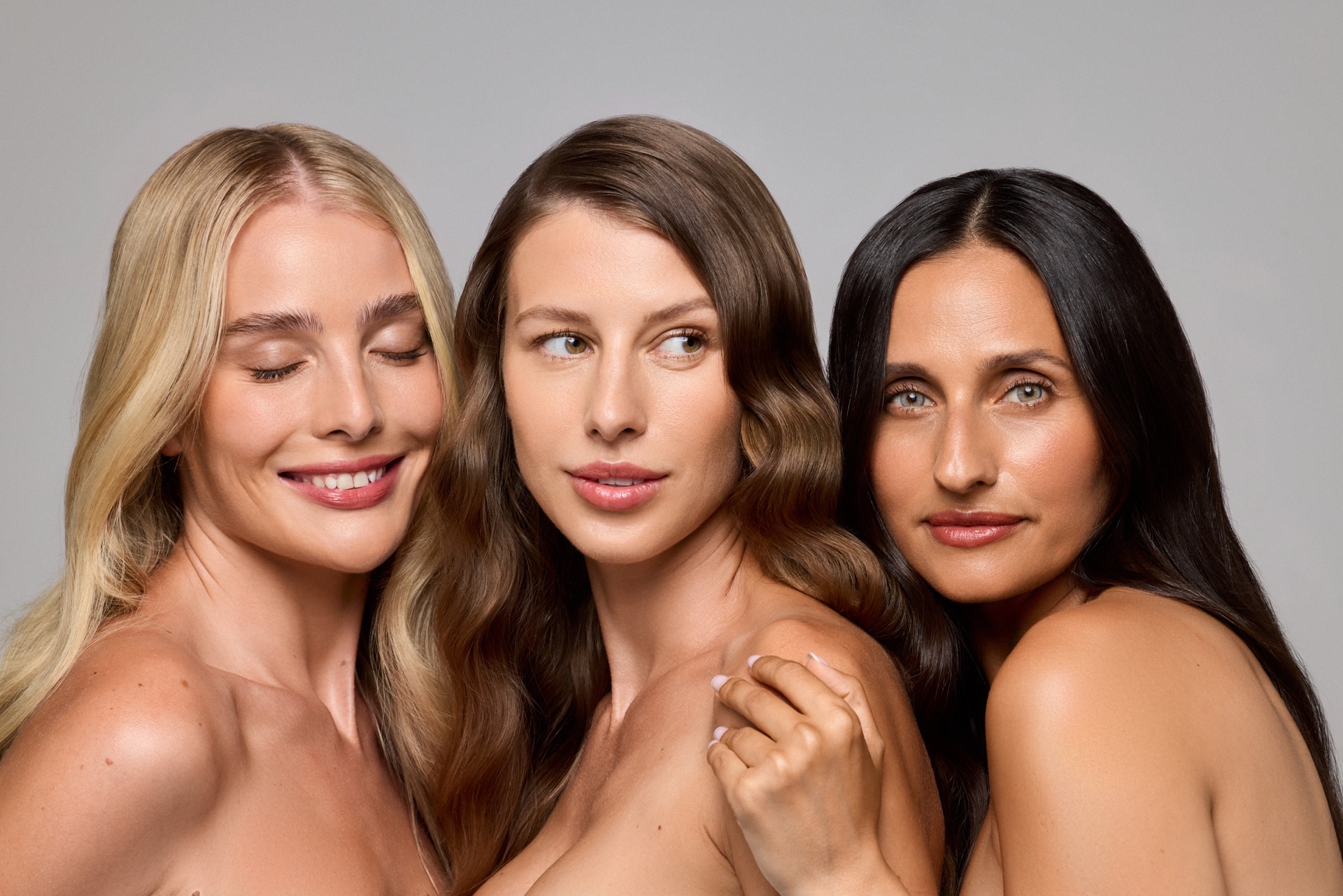 Three models with long, glossy hair — blonde, brunette, and black — posed close together against a soft grey backdrop, one resting her hand on another's shoulder.