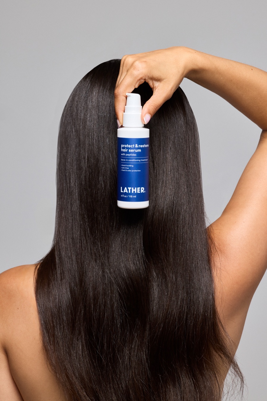 Seen from behind, a model holds a bottle of LATHER Protect & Restore Hair Serum against her long, straight black hair.