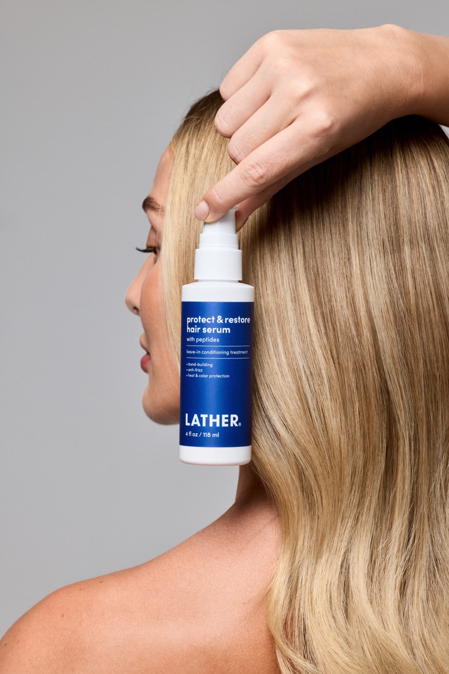 A model in profile holds a bottle of LATHER Protect & Restore Hair Serum against her sleek blonde hair, one hand lifting a section from her crown.