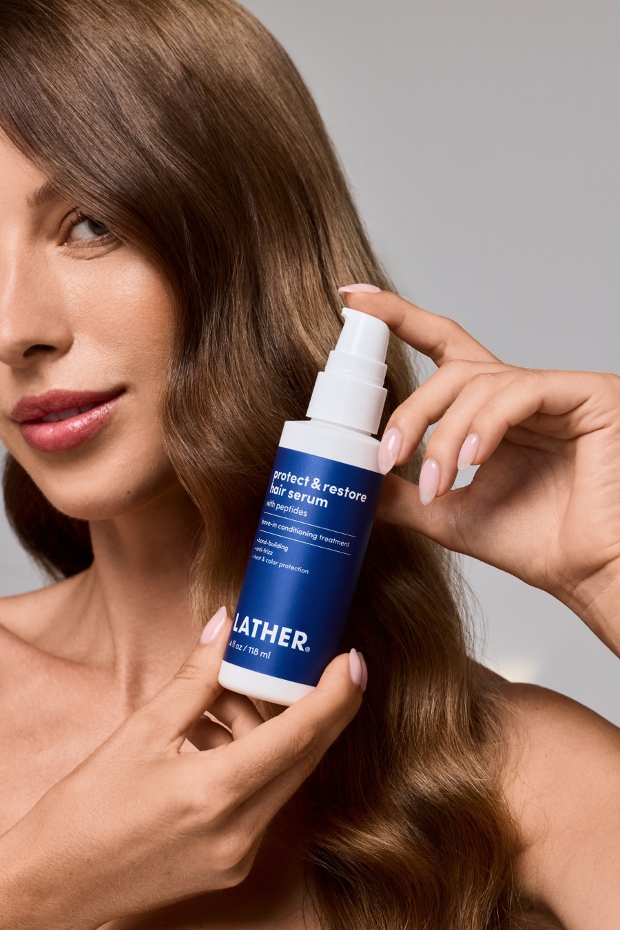Close-up of a model with glossy brown waves holding a bottle of LATHER Protect & Restore Hair Serum beside her face.