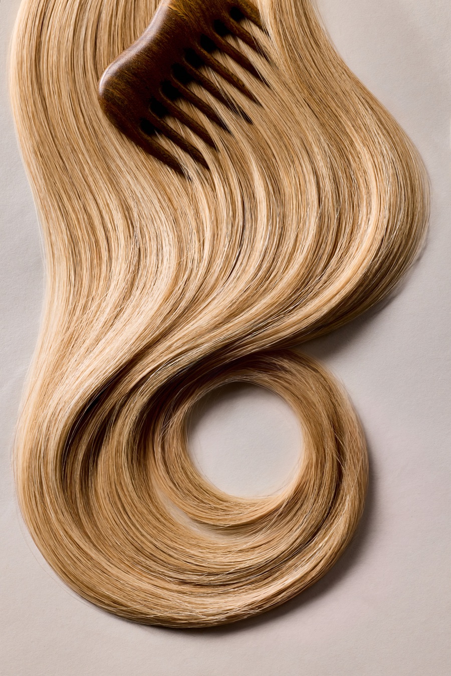 A long sweep of glossy honey-blonde hair curling into a loop on a pale backdrop, a wooden comb resting at the top.