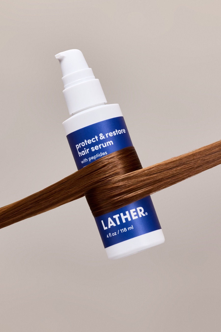 A bottle of LATHER Protect & Restore Hair Serum tilted on a warm grey backdrop, a strand of glossy auburn hair wrapped around its middle.