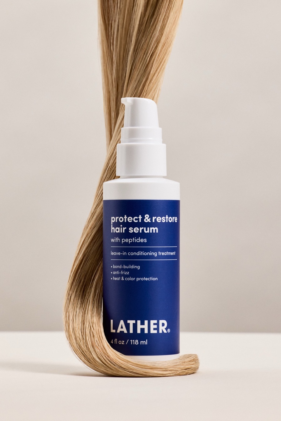 A bottle of LATHER Protect & Restore Hair Serum standing on a pale surface, a long strand of honey-blonde hair looping down behind it and curling around its base.
