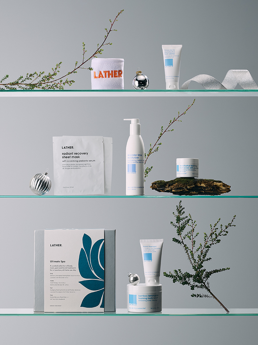 The LATHER Ultimate Spa gift box shown with its contents — a monogrammed LATHER towel, lavender lime moisturizer, eucalyptus foaming foot scrub, hand therapy cream, and a radiant recovery sheet mask — styled with silver ornaments and greenery.