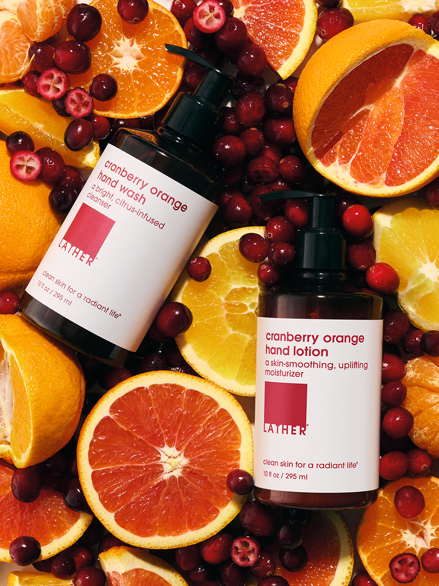 LATHER Cranberry Orange hand wash and hand lotion bottles nestled among sliced oranges and scattered fresh cranberries.