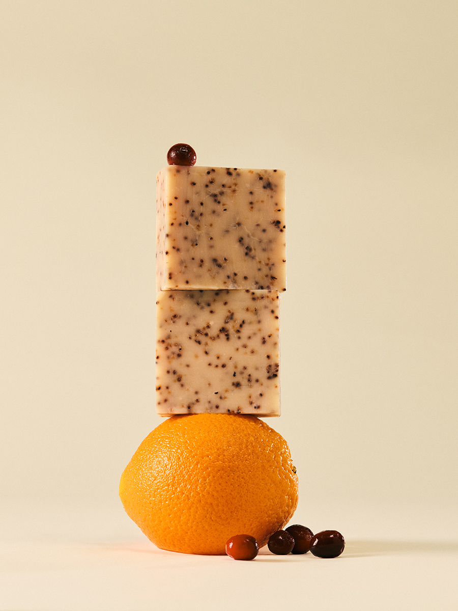 Two speckled LATHER soap bars stacked and balanced on top of a whole orange, with loose cranberries scattered at its base.