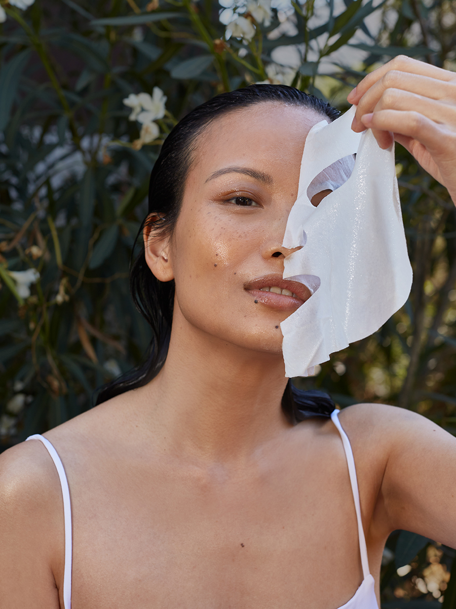 A model outdoors holds a white sheet mask half-covering her face amid green foliage, one eye and freckled cheek visible.