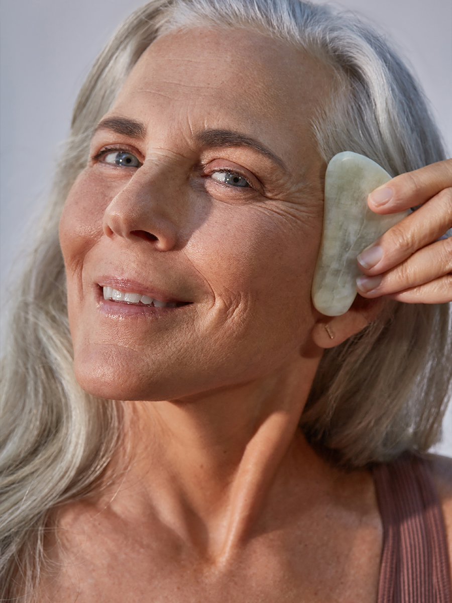 Portrait of a silver-haired older woman smiling and holding a jade gua sha stone to her cheek.