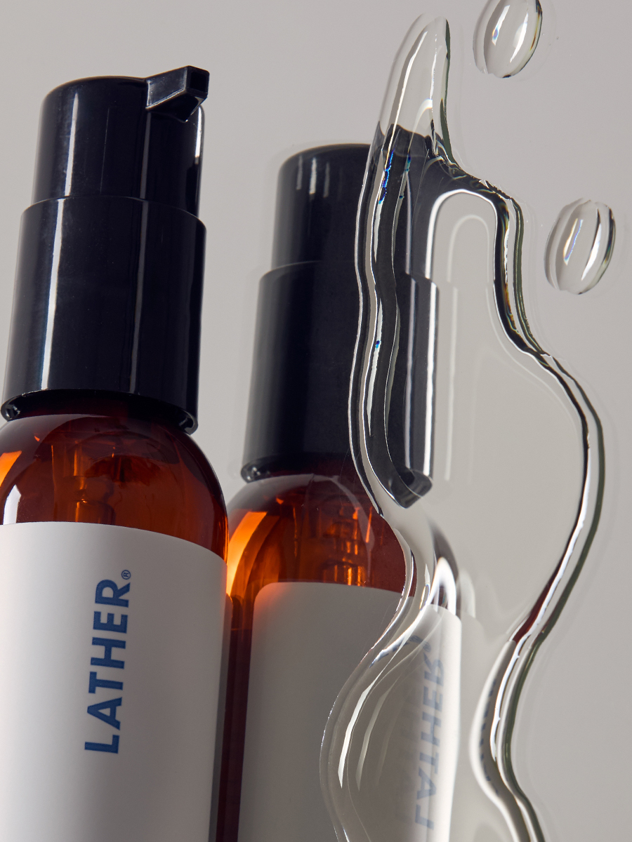 Two amber Lather pump bottles with clear liquid splashing and pooling between them.