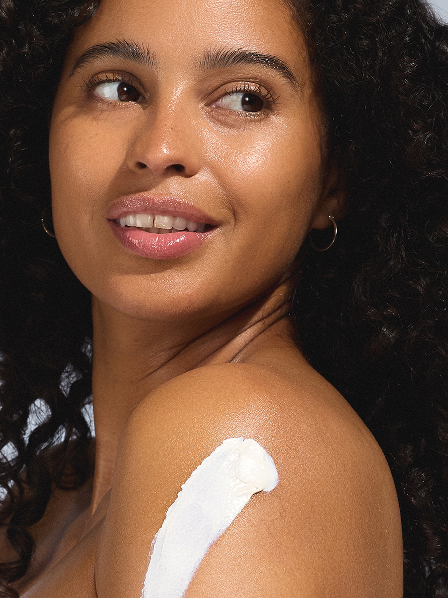 A model glances over her bare shoulder with a swipe of white sunscreen applied, dark curly hair falling around her face.