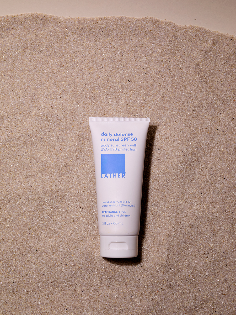 A tube of Lather daily defense mineral SPF 50 lying flat on a sandy beach background.