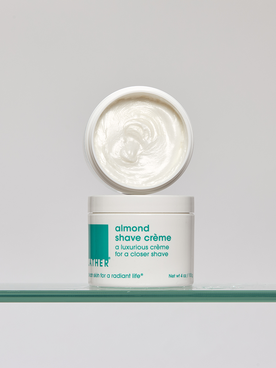 An open jar of Lather almond shave crème resting on a glass shelf, its lid tilted to reveal the whipped texture inside.