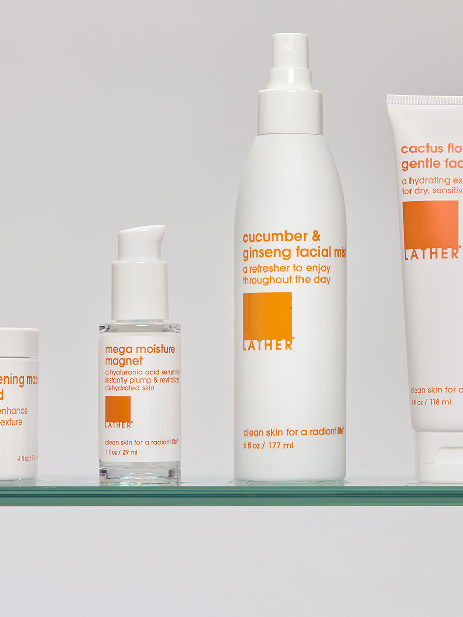 A row of Lather products — mega moisture magnet serum, cucumber & ginseng facial mist, cactus flower & aloe scrub, and a moisture mask — lined up on a glass shelf.