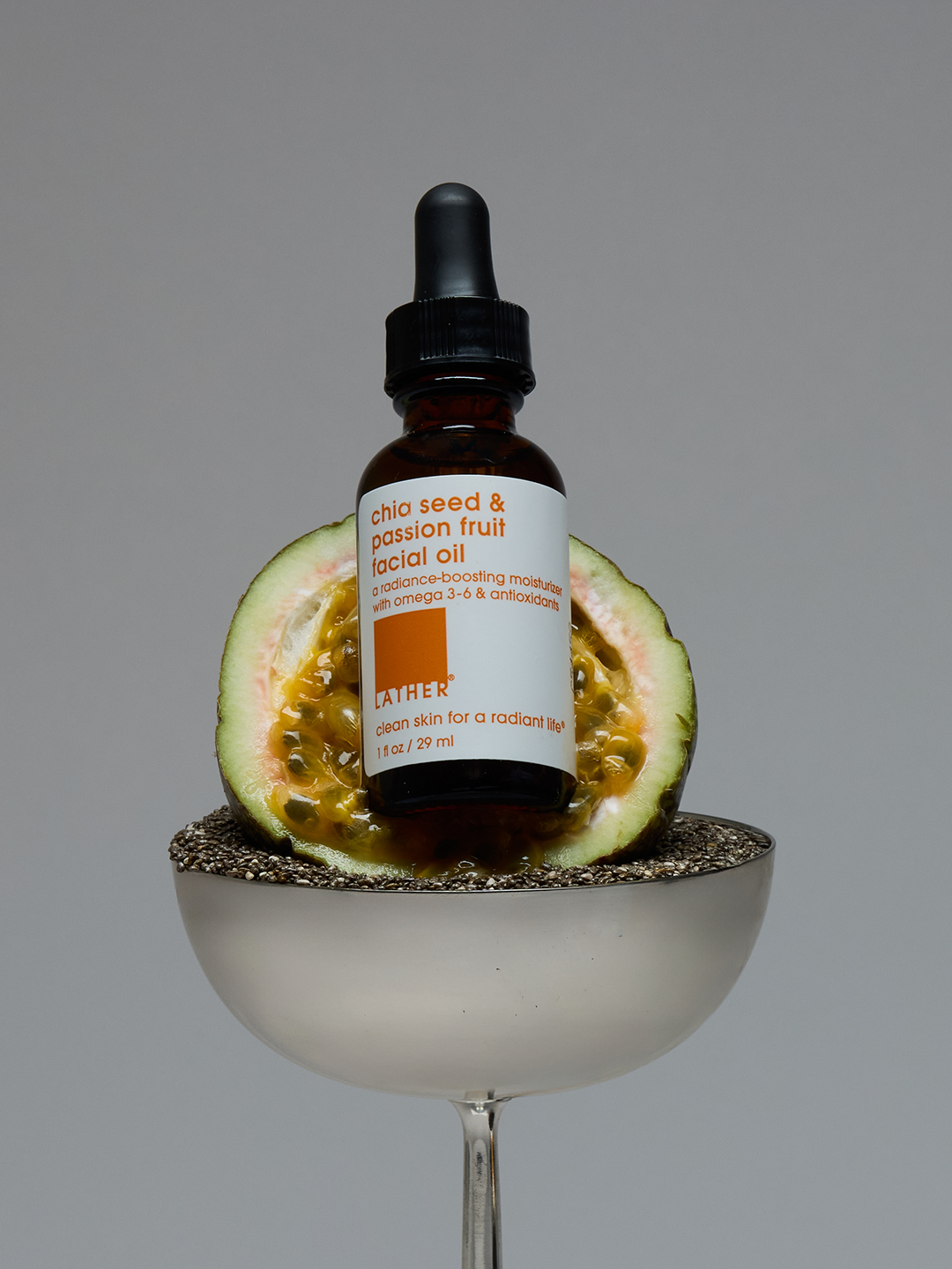 Lather Chia Seed & Passion Fruit Facial Oil resting against a halved passion fruit on a bed of chia seeds in a silver coupe glass.
