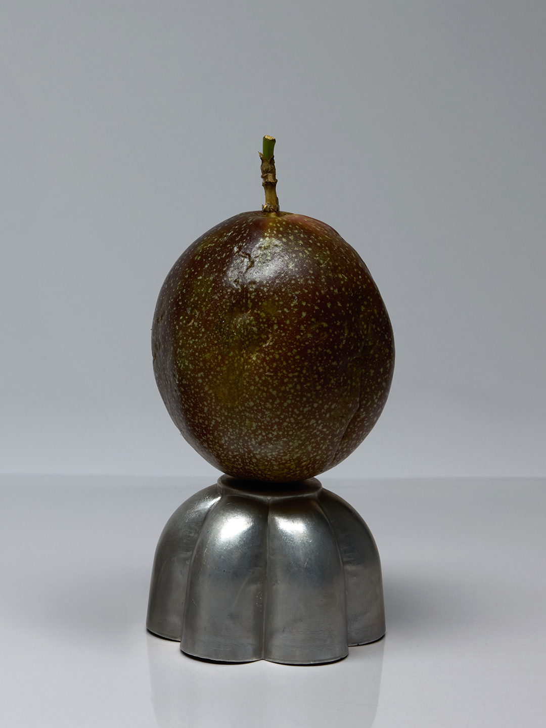 Lather Chia Seed & Passion Fruit Cleansing Oil bottle beside a whole passion fruit, both resting on pewter stands.