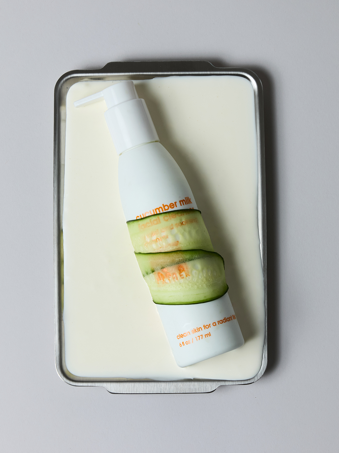 Overhead view of Lather Cucumber Milk Facial Cleanser wrapped in cucumber slices, resting on a milk-glazed metal tray.