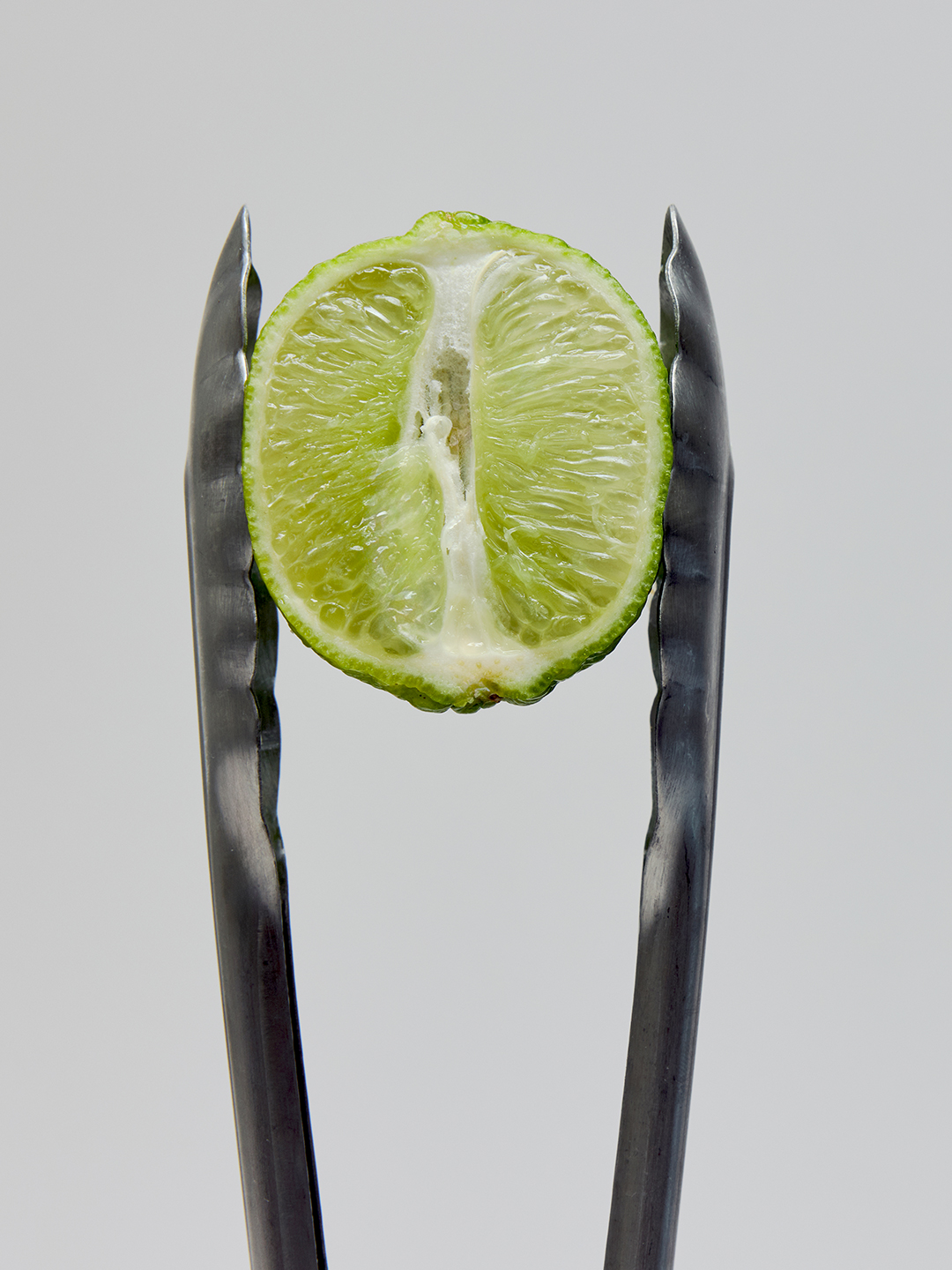 A halved kaffir lime held upright between the tines of black metal tongs.