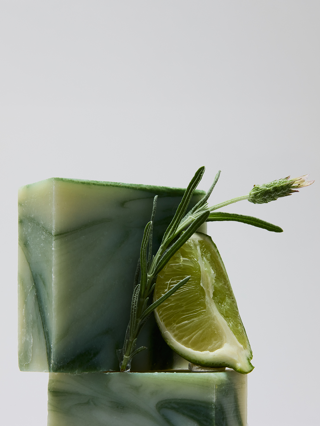 A green marbled Lather soap bar accented with a lime wedge, a rosemary sprig, and a lavender sprig.