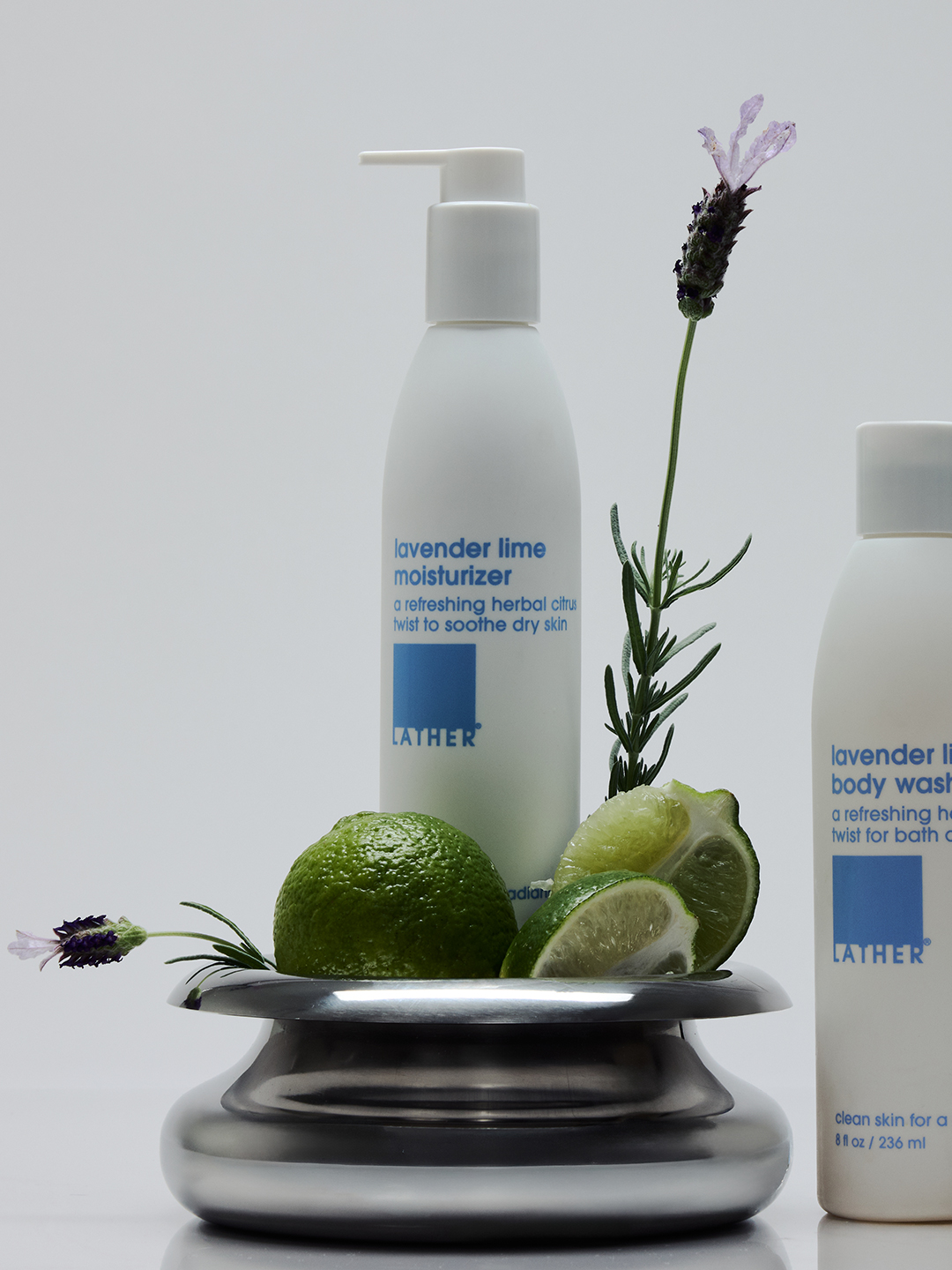 Lather Lavender Lime Moisturizer and Body Wash bottles surrounded by fresh limes, rosemary, and lavender sprigs on a metal pedestal dish.