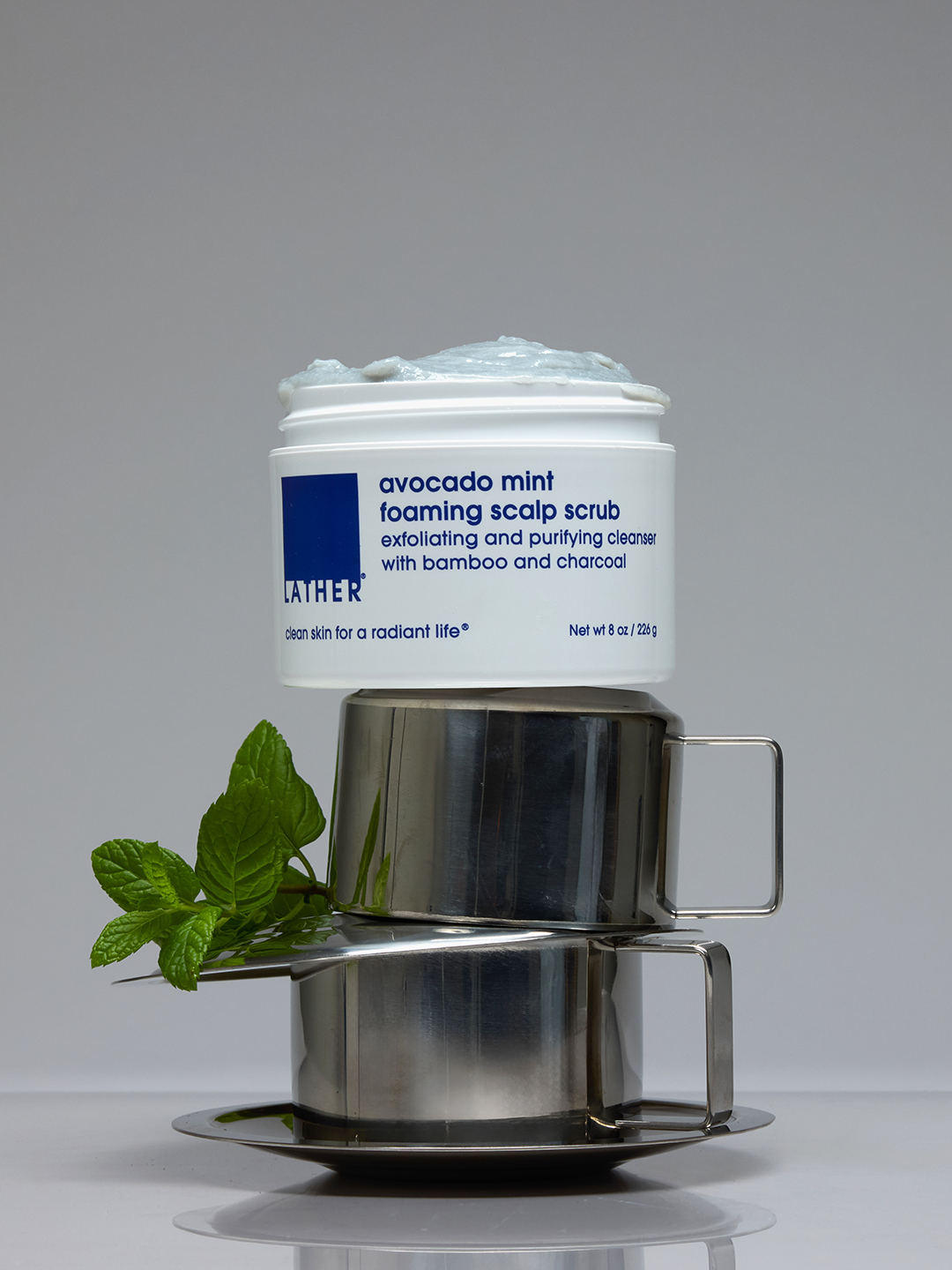 Lather Avocado Mint Foaming Scalp Scrub resting atop stacked metal cups with a fresh mint sprig, on a round metal tray.